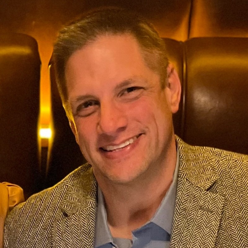 Portrait of a smiling man in a herringbone blazer in a warmly lit lounge setting on brown leather seats.