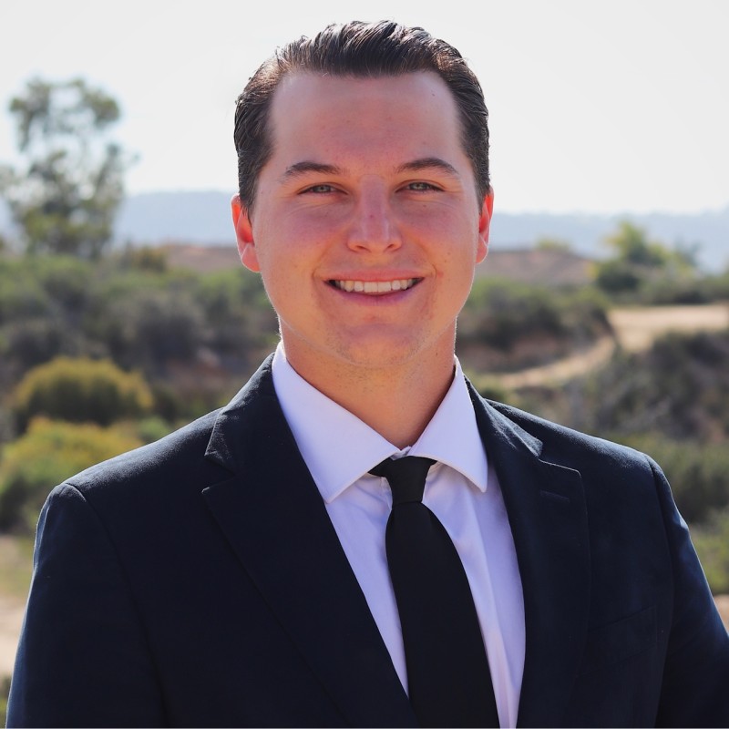 Professional head-and-shoulders portrait of a smiling man in a black suit outdoors.