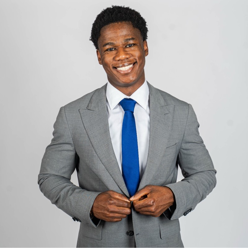 Smiling man in a gray suit adjusting his jacket and blue tie against a plain background