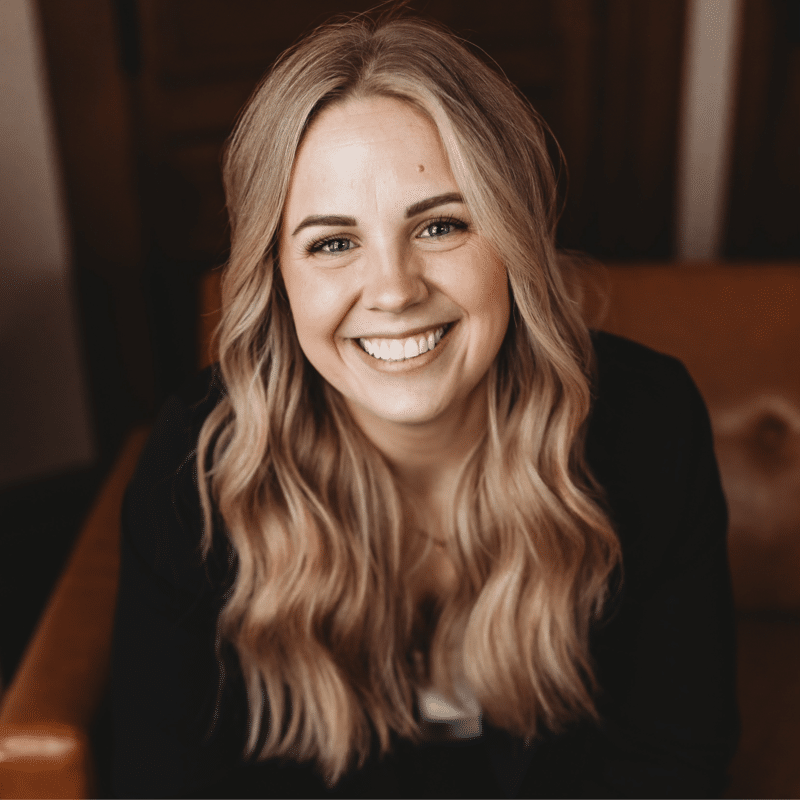 Smiling woman with long wavy blond hair in a black top, indoors, facing the camera.