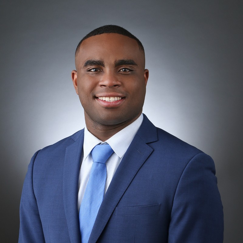 Smiling man in a navy suit and light blue tie, posing for a professional headshot against a gray studio backdrop.