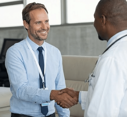 Two men shake hands in a medical office lobby; one wears a stethoscope, indicating a healthcare setting.