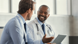 Two doctors in a clinical setting share a friendly discussion, one holding a tablet while smiling at the other with a stethoscope around his neck.