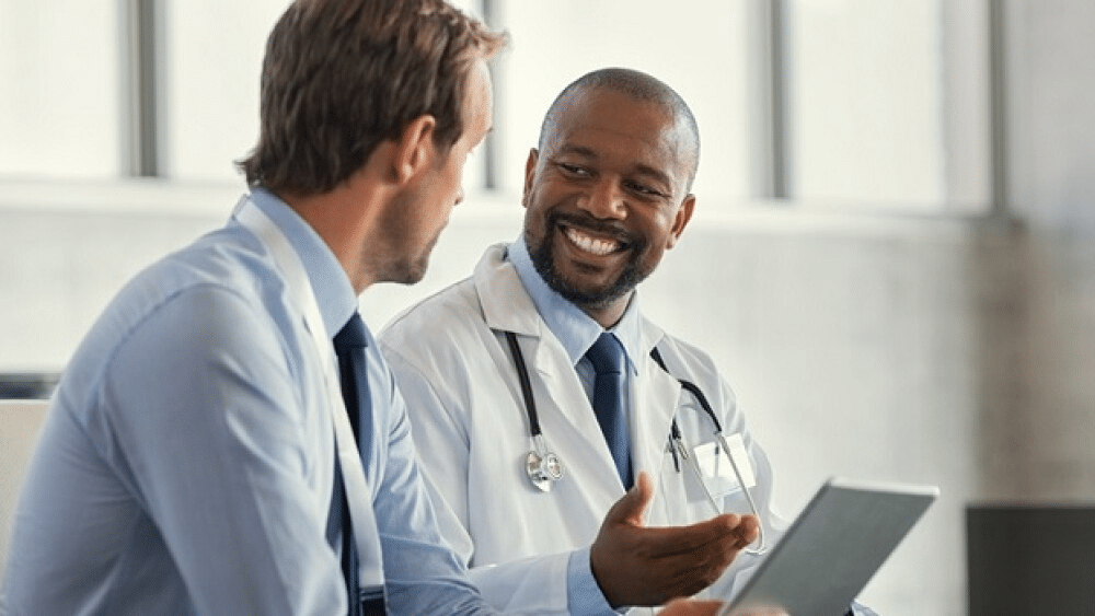 Two doctors in a clinical setting share a friendly discussion, one holding a tablet while smiling at the other with a stethoscope around his neck.