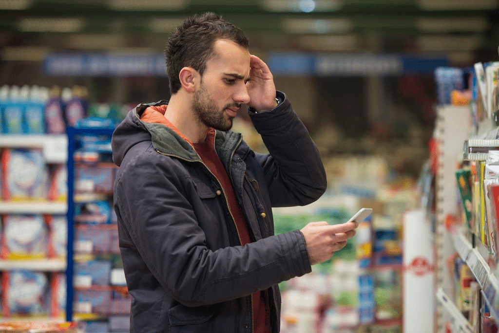 Man in a dark jacket in a store checks his phone, hand to forehead in a puzzled pose.
