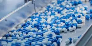 Blue and white capsules on a metal conveyor belt in a pharmaceutical manufacturing line.