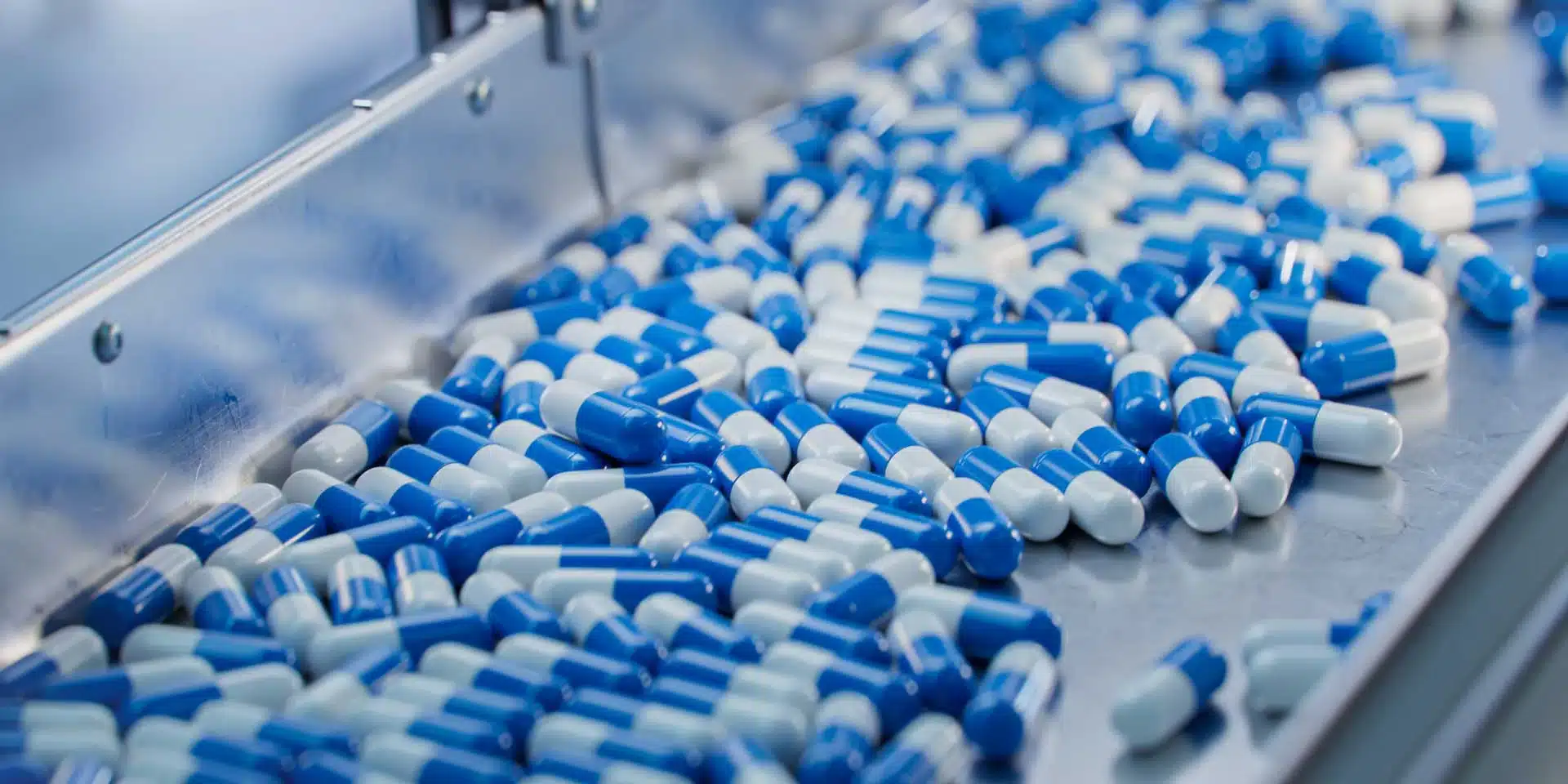 Blue and white capsules on a metal conveyor belt in a pharmaceutical manufacturing line.