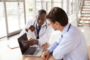 Two doctors sit at a bright clinic desk, one points at a tablet while the other types on a laptop with a stethoscope nearby.