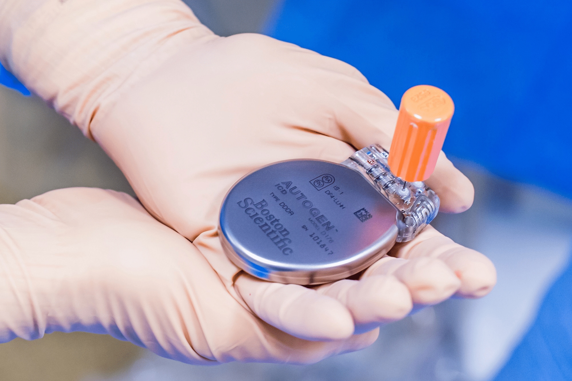 Gloved hands holding a small medical device with an orange cap in a clinical setting