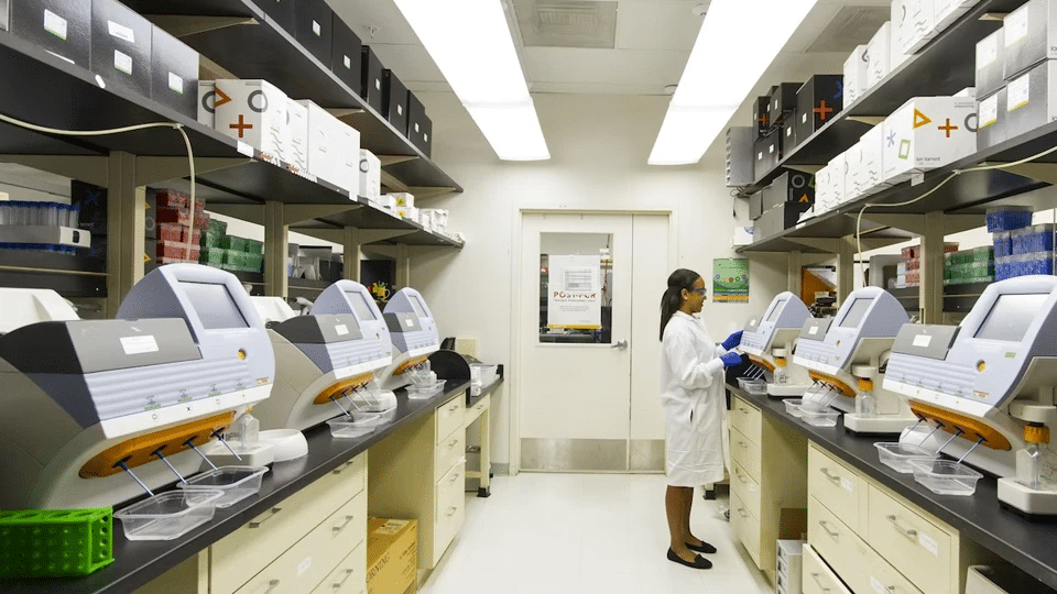 Laboratory scene: a technician in a white coat and gloves operates automated analyzers along benches with shelves full of boxes.