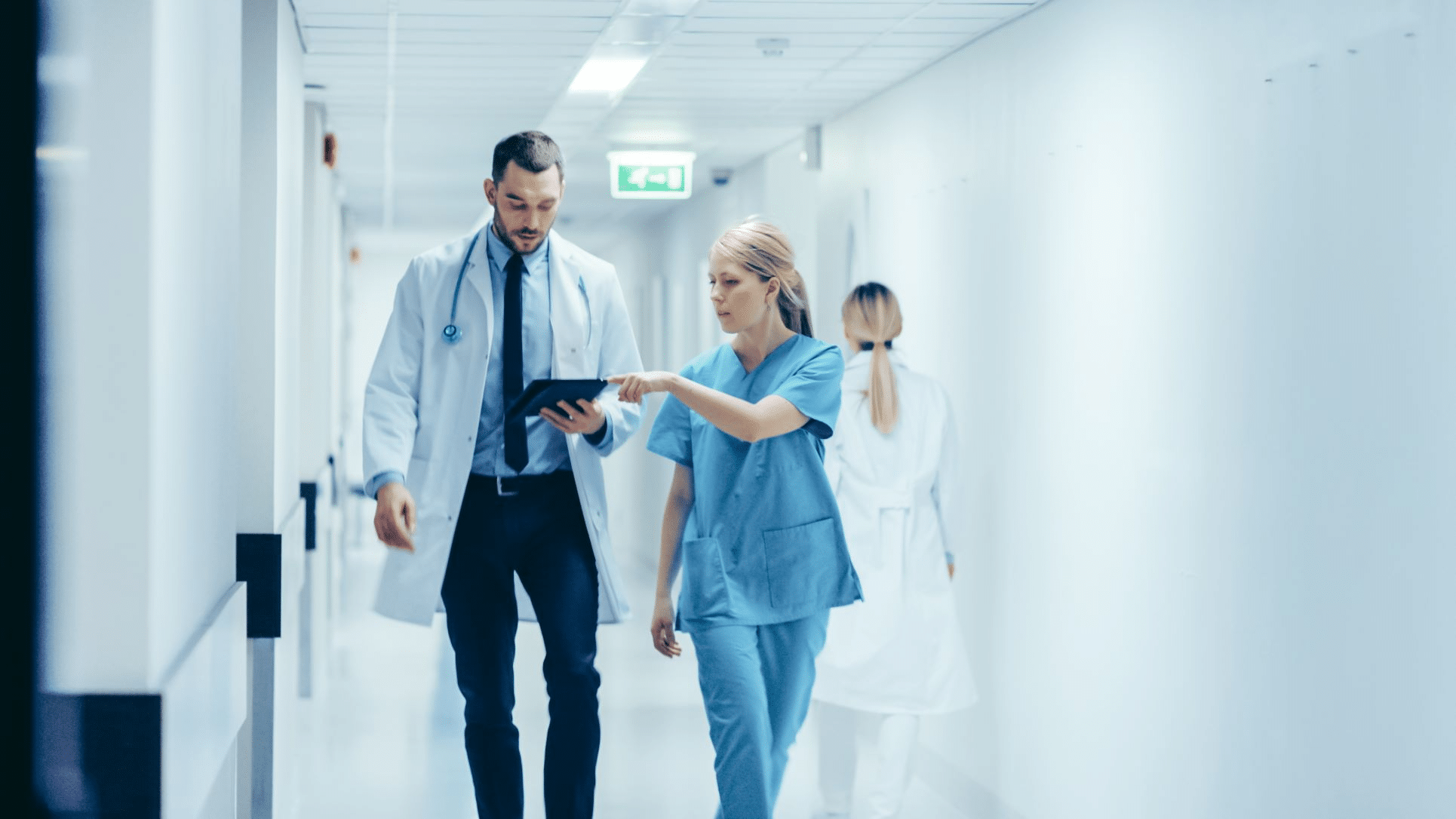 Doctor in a white coat and stethoscope reviews a tablet with a nurse in blue scrubs as they walk down a bright hospital hallway, collaborating.